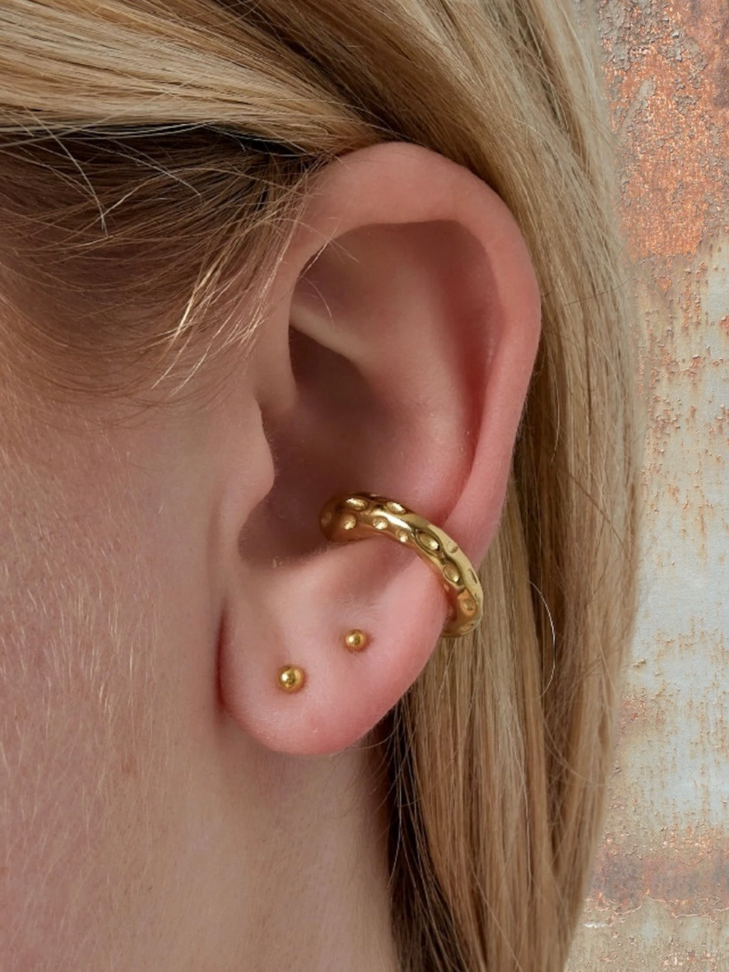 WILDSIDE gold-tone metal ear cuff with textured design, no piercing needed. Worn with two small gold stud earrings.